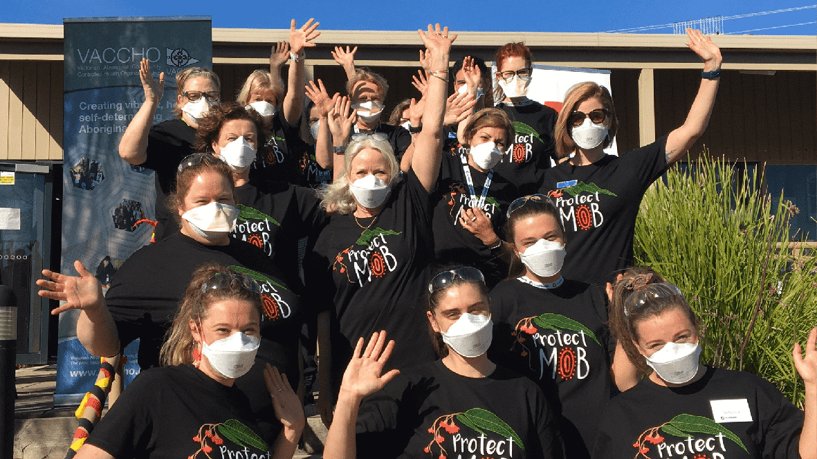 A large group of health professionals wearing masks and black t-shirts with the words "Protect Mob" on the front waving to the camera.
